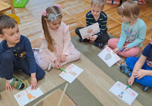 Dzieci siedząc na dywanie zaznaczają klamerką na kartach demonstracyjnych zmysł odpowiadający obrazkowi.