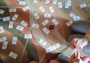 Pomoce dydaktyczne leżące na dywanie, które przedstawiają kawałki pizzy z różną ilością wędliny oraz kartoniki z cyframi, które dzieci odpowiednio ze sobą łączą.