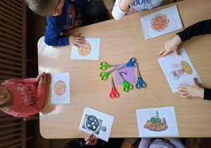 Dzieci siedzą przy stoliku i przygotowują się do ułożenia obrazków przedstawiających rzeczy związane z pizzą, które najpierw muszą rozciąć za pomocą nożyczek.