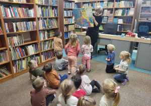 Dzieci siedzą na dywanie, pani bibliotekarka pokazuje wielką książkę.