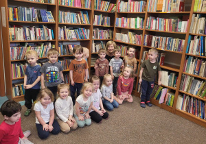 Dzieci wraz z panią bibliotekarką pozują do zdjęcia na tle regałów wypełnionych książkami.