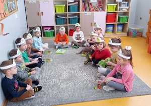 Przedszkolaki siedzą na dywanie i prezentują zabawki i książki związane z dinozaurami.