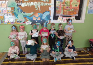 Dzieci z grupy Krasnale prezentują się w swoich dinowych maskach oraz pokazują dyplomy za udział w przygodach z dinozaurami.