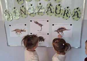 Dwie dziewczynki oglądają obrazki dinozaurów na tablicy tematycznej.