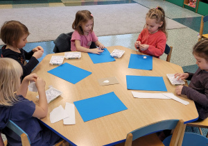 Dziewczynki siedzą przy stoliku i wyklejają szablon zęba wydzieranką z białego papieru.