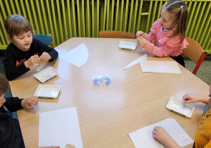 Dziewczynki i chłopcy siedzą przy stoliku i wydzierają białą kartkę na małe kawałki.