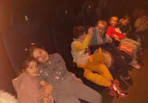 Dzieci z grupy Tropiciele siedzą w fotelach kinowych i czekają na bajkę pt. „Zagadka Klary Muu”.