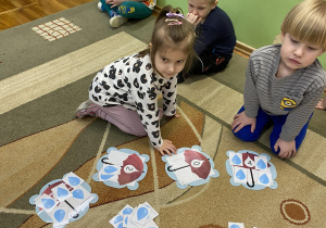 Dzieci siedzą na dywanie, dwoje dzieci odczytuje cyfrę z wybranego obrazka parasola i dokłada odpowiednią liczbę kropek.