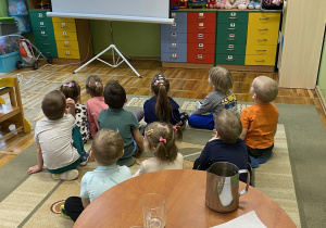 Dzieci siedzą na dywanie i oglądają film edukacyjny pt. „W marcu jak w garncu”.