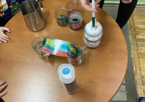 Dzieci stoją przy stoliku i razem z nauczycielką wykonują eksperyment „Kolorowa chmurka”.
