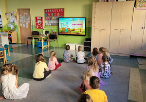 Dzieci z grupy Motylki siedzą na dywanie i oglądają filmik edukacyjny o Dniu Kobiet.