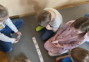 Dzieci siedzą na dywanie i grają w matematyczne domino.