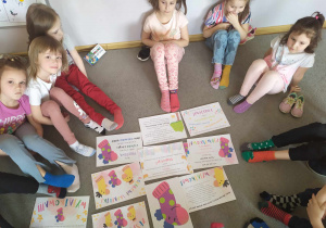 Dzieci siedzą na dywanie i słuchają bajki „Wyjątkowa skarpetka” oglądając ilustarcje do czytanej bajki.
