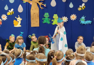 Przedszkolanka w pelerynie z kroplami i z butelką z wodą dłoni przedstawi swoją rolę.