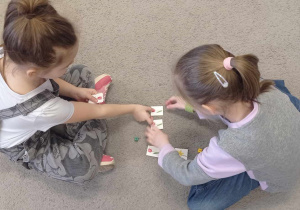 Zdjęcie przedstawia dwie dziewczynki podczas zabawy matematycznej z kostką i fiszkami.
