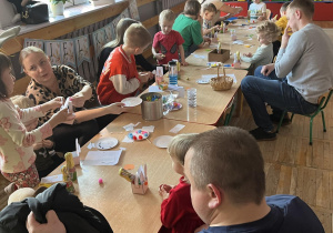 Dzieci z rodzicami siedzą przy stolikach, wykonują papierowe koszyczki.