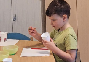 Chłopiec siedzi przy stoliku i maluje farbą papierowy kubek.