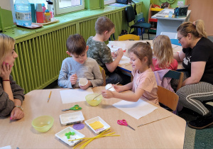 Dziewczynka i chłopiec siedzą przy stoliku i z pomocą Pani malują papierowe kubeczki farbą.