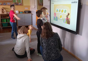 Zdjęcie przedstawia dzieci stojące przed monitorem wraz ze swoimi rodzicami, grających w grę „ Co nie pasuje”.