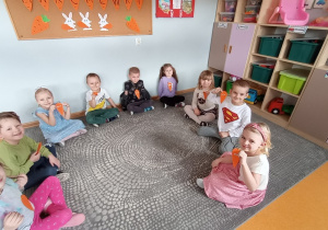 Przedszkolaki siedzą w kole na dywanie i prezentują marchewkowe medale.