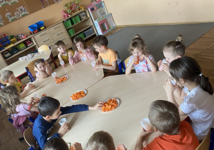 Dzieci siedzą przy stoliku i degustują sok marchwiowy oraz chrupią surową marchewkę.