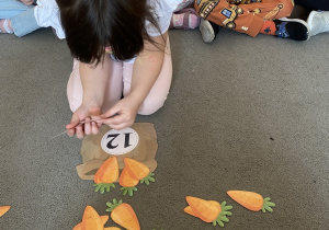 Dziewczynka siedzi na dywanie i układa tyle marchewek ile wskazuje liczba na worku.