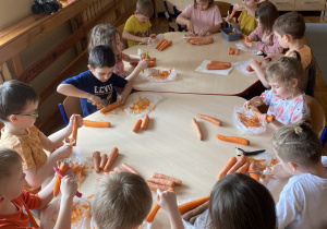 Przedszkolaki siedzą przy stoliku i samodzielnie obierają marchewki używając obieraczki.