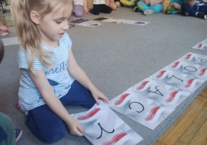 Dziewczynka w bluzce w paski układa na dywanie literę do napisu Słynni Polacy.