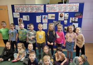 Dzieci z grupy Żabki pozują do zdjęcia na holu przedszkolnym przed tablicą z wystawą Słynni Polacy.