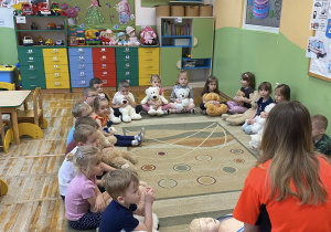 Dzieci siedzą na dywanie z maskotkami i słuchają Pani ratownik, która opisuje procedurę udzielania pierwszej pomocy.