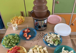 Na stoliku stoi fontanna czekoladowa oraz na talerzykach owoce.