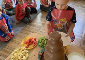 Chłopiec macza owoc w czekoladzie reszta dzieci siedzi na dywanie.