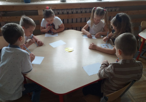 Dzieci ćwiczą aparat artykulacyjny przenosząc papierowe kostki czekolady na kartkę papieru za pomocą słomki.