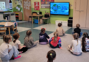 Dziewczynki i chłopcy siedzą na dywanie i oglądają filmik edukacyjny o Ziemi.