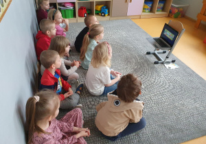 Dzieci siedzą na dywanie i oglądają film edukacyjny „Dobre rady na złe odpady”.