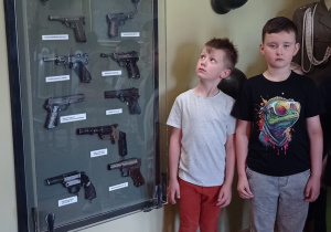 Zdjęcie przedstawia dwóch chłopców pozujących do zdjęcia przy gablocie z pistoletami.