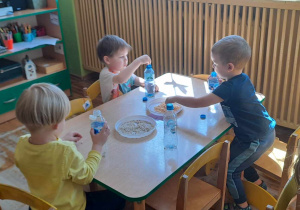 Chłopcy z siedząc przy stoliczku wykonują ,,grzechotki” z butelek plastikowych i sypkich produktów spożywczych.