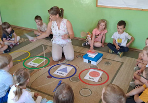 Dzieci siedząc na dywanie oglądają plansze z figurami oraz pudełko sensoryczne.