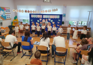Dzieci stoją przy tablicy z dekoracją i śpiewają piosenkę dla rodziców. W tle siedzący na krzesłach rodzice.