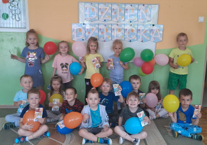 Dzieci z grupy Misie pozują do zdjęć na tle tablicy dekoracyjnej. W rękach trzymają balony oraz upominki.