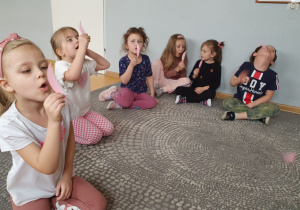 Dzieci siedzą na dywanie wykonują ćwiczenia oddechowe dmuchając na różowe piórka.
