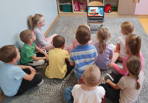 Dzieci siedzą na dywanie oglądają film edukacyjny dotyczący bezpieczeństwa w sieci.