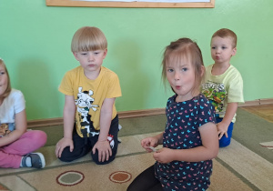Dzieci siedzą na dywanie i wykonują ćwiczenia artykulacyjne.