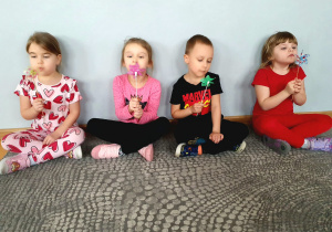 Dzieci siedzą na dywanie wykonują ćwiczenia oddechowe z wykorzystanie wiatraczków.