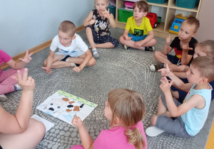 Przedszkolaki siedzą w kole na dywanie i rozwiązują zadania matematyczne na podstawie prezentowanego obrazka.