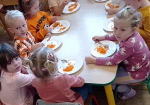 Zdjęcie przedstawia grupę dzieci siedzącą przy stoliku podczas degustacji marchewkowej surówki.