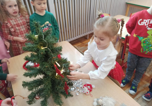 Dziewczynka z grupy Słoneczka wiesza kolorową bombkę na przedszkolną choinkę.