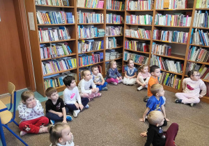 Dzieci siedzą na dywanie w bibliotece, słuchają ciekawostek na temat miejsca w którym się znajdują.