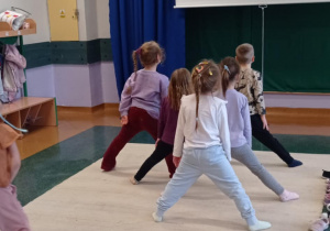 Zdjęcie przedstawia grupę dzieci wykonującą ćwiczenia jogi. Przedszkolaki stoją w rozkroku.