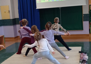 Zdjęcie przedstawia grupę dzieci wykonującą ćwiczenia jogi. Przedszkolaki stoją w rozkroku z rękoma wyprostowanymi w bok.